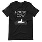 Front of a black heather t-shirt that reads "House Cow" in white text over an illustration that is the white silhouette of a large dog laying on the dark silhouette of a couch.